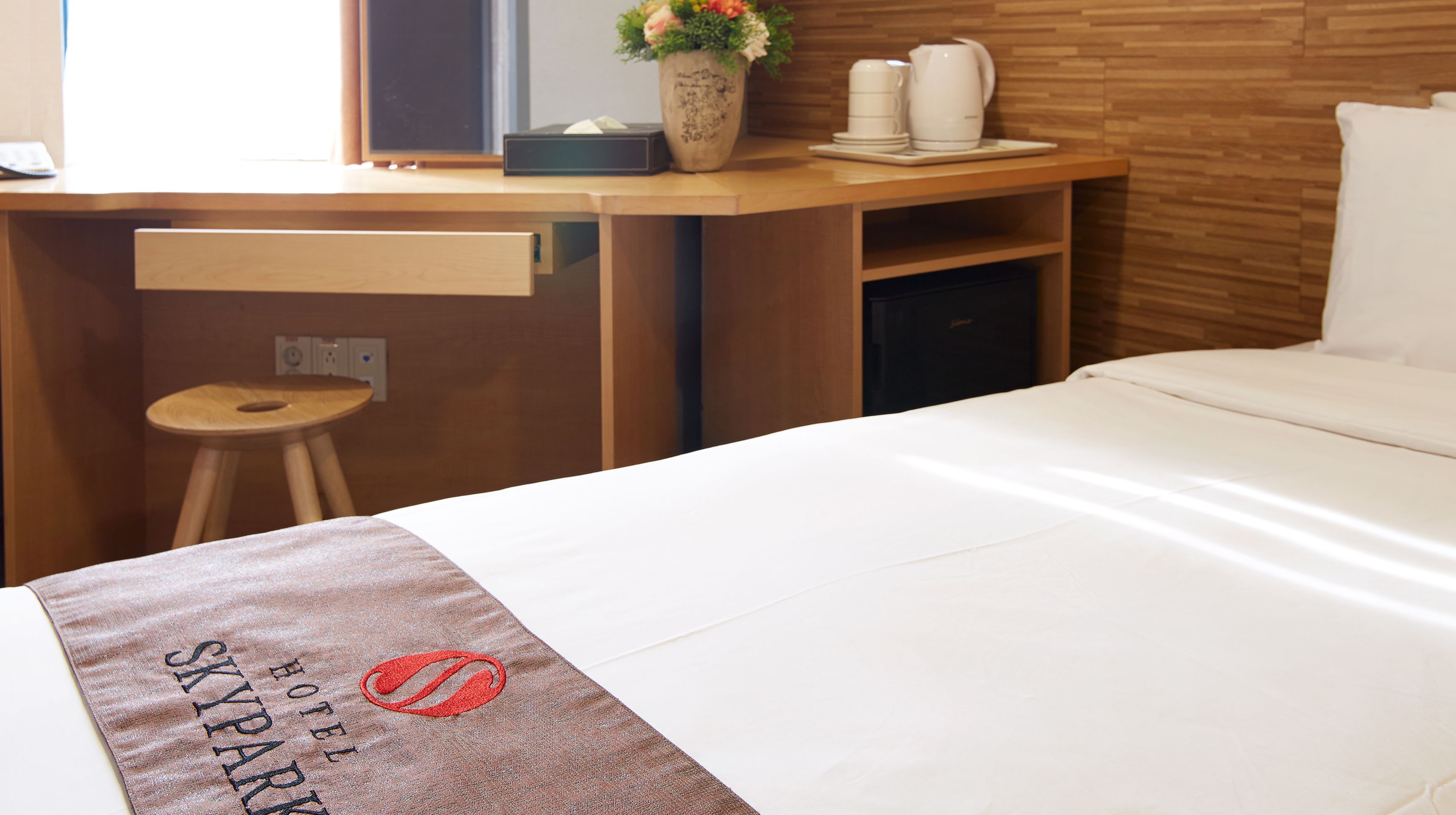 A hotel room with a bed, a desk, a chair, a small stool, a vase with flowers, and a coffee maker.