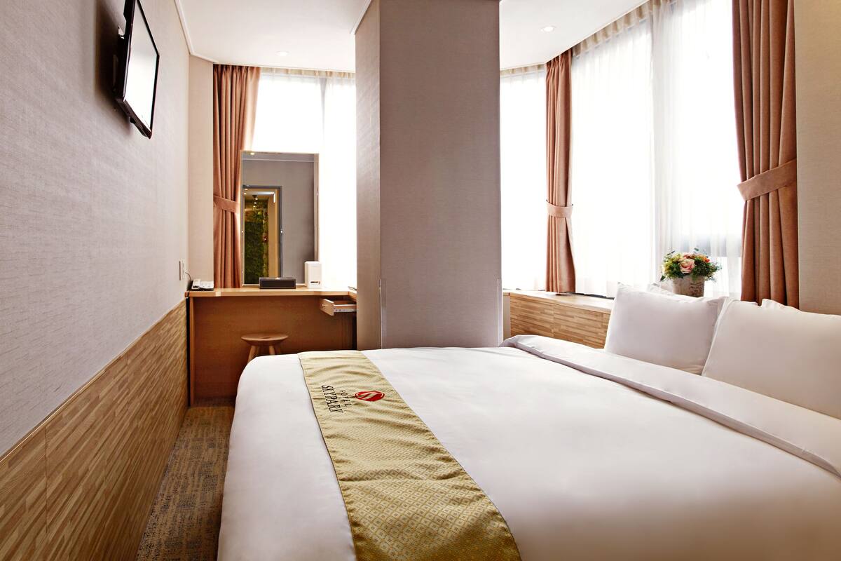 A hotel room with a large bed, a desk, a chair, a mirror, and a window with curtains.