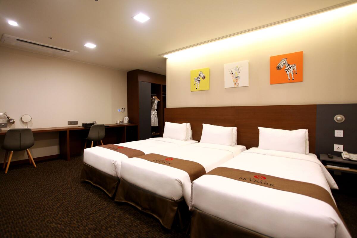 A hotel room with two beds, a desk, and chairs. There are wall art pieces and a coat rack.