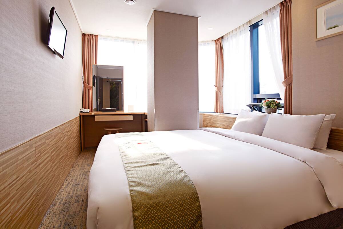 A hotel room with a large bed, a desk with a chair, a mirror, and a window with curtains.
