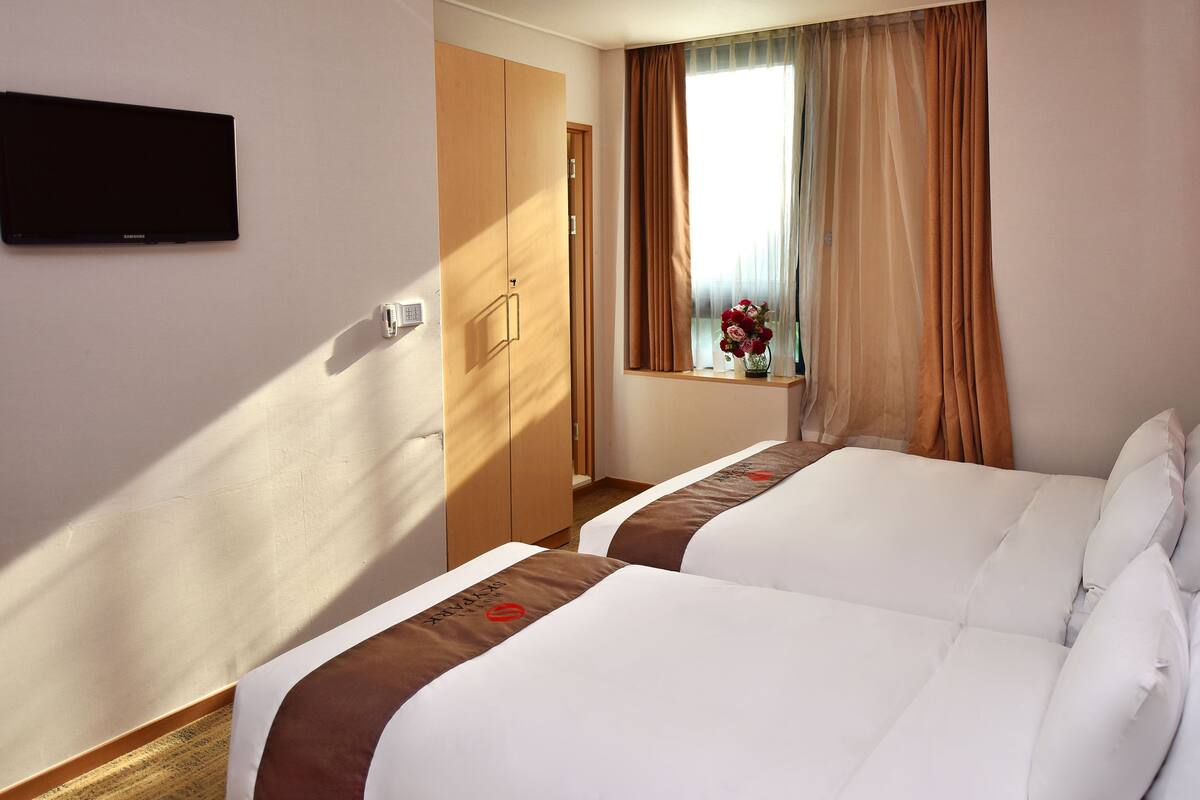 A hotel room with two beds, a window with curtains, a bedside table with a vase of flowers, and a wall-mounted TV.