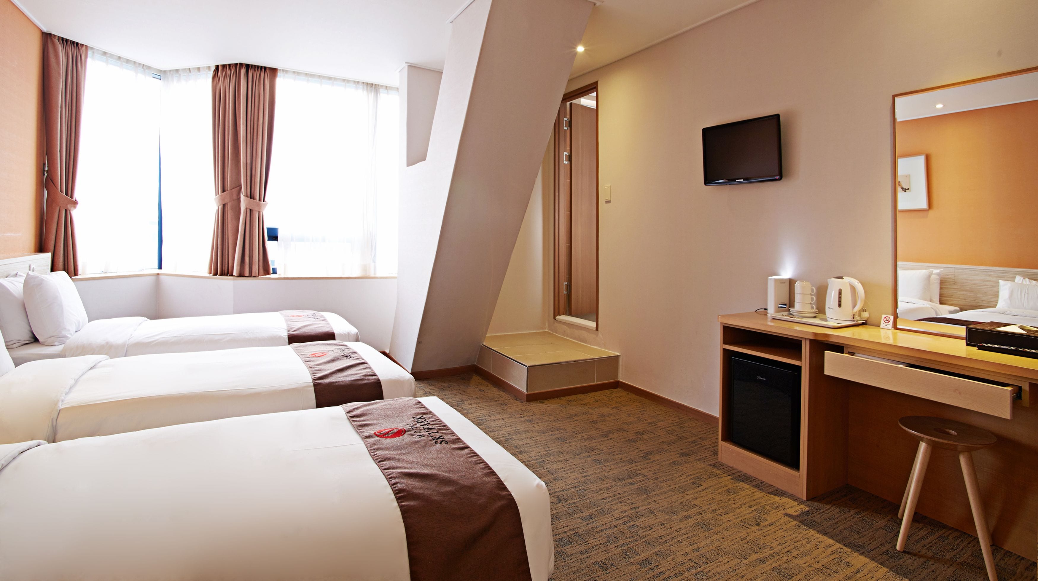 A hotel room with two beds, a desk with a coffee maker, a TV, and a shower area.