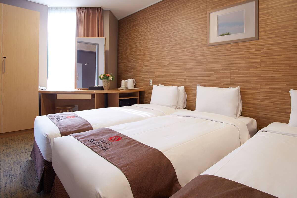 A hotel room with two beds, a desk, a chair, a coffee maker, and a vase with flowers.