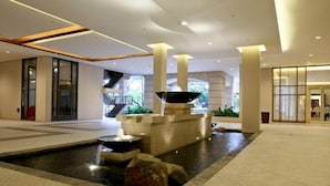 A modern indoor lobby with a central water feature, large pillars, and a staircase leading to upper levels. - Joya Lofts and Towers (Makati)