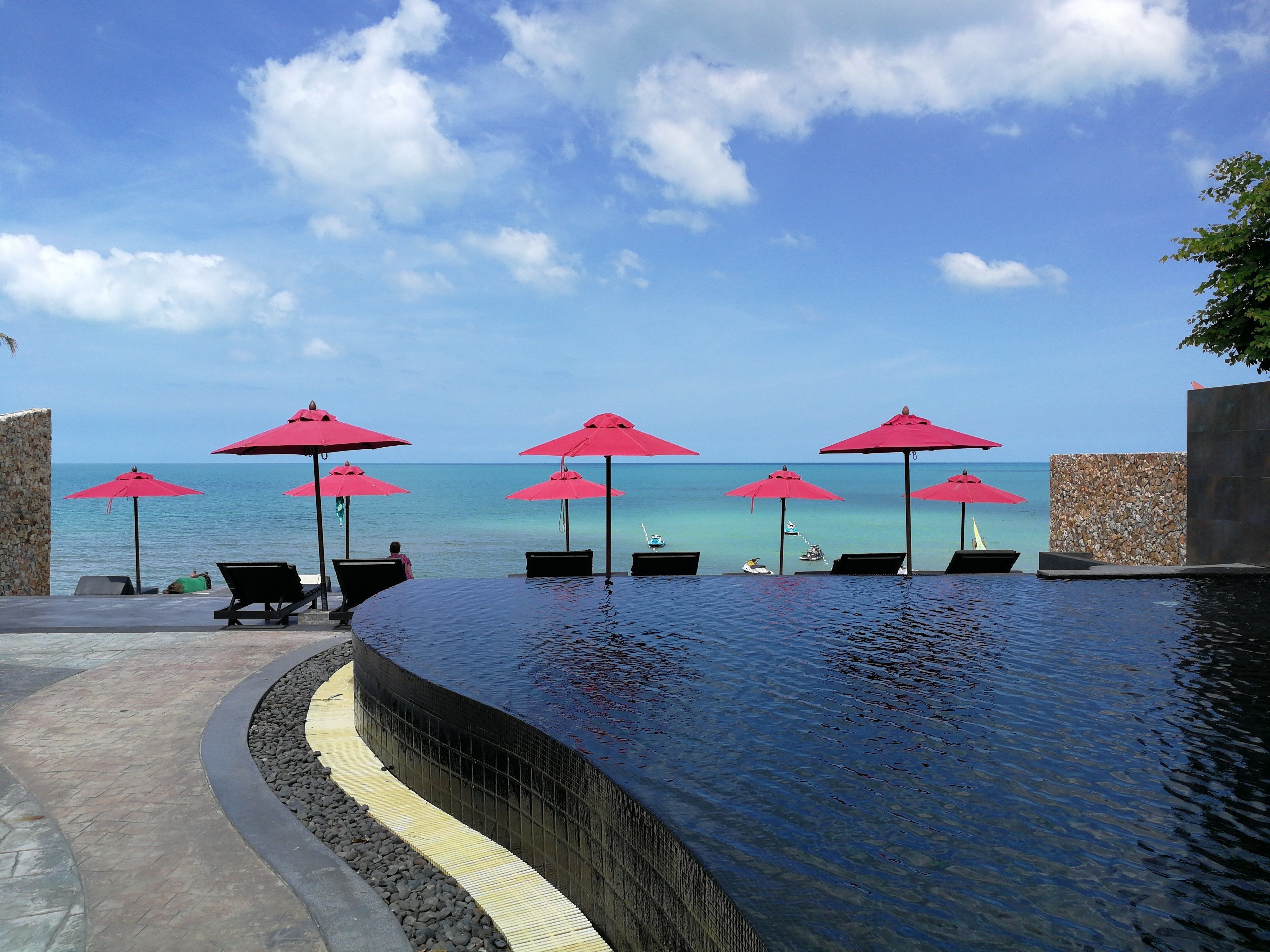 A poolside area with lounge chairs and red umbrellas, overlooking a clear blue sea.