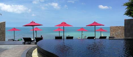 A poolside area with lounge chairs and red umbrellas, overlooking a clear blue sea.
