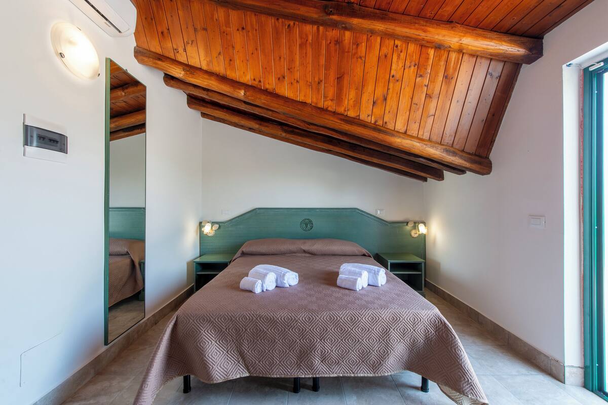 A bedroom with a wooden ceiling, a bed with a patterned bedspread, two bedside lamps, and a mirror on the wall.