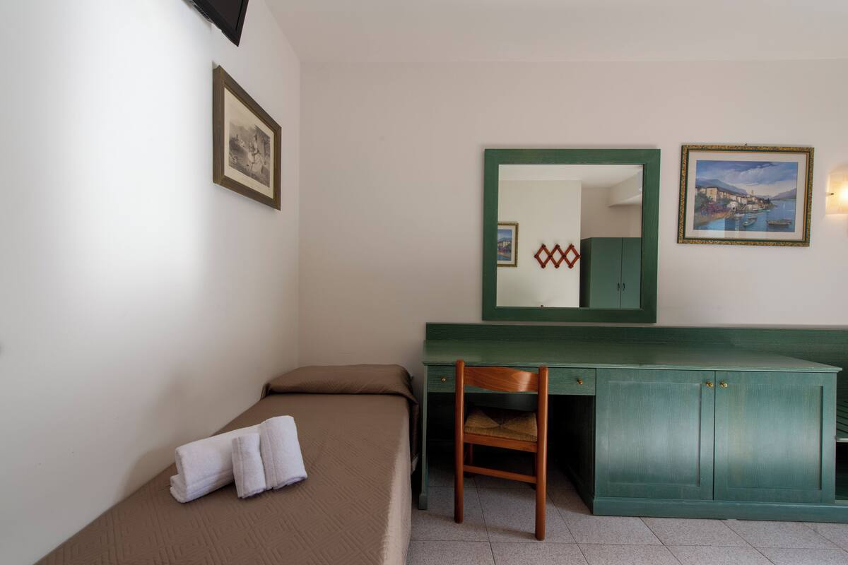A small, clean room with a bed, a desk, and a chair. There are two framed pictures on the wall.