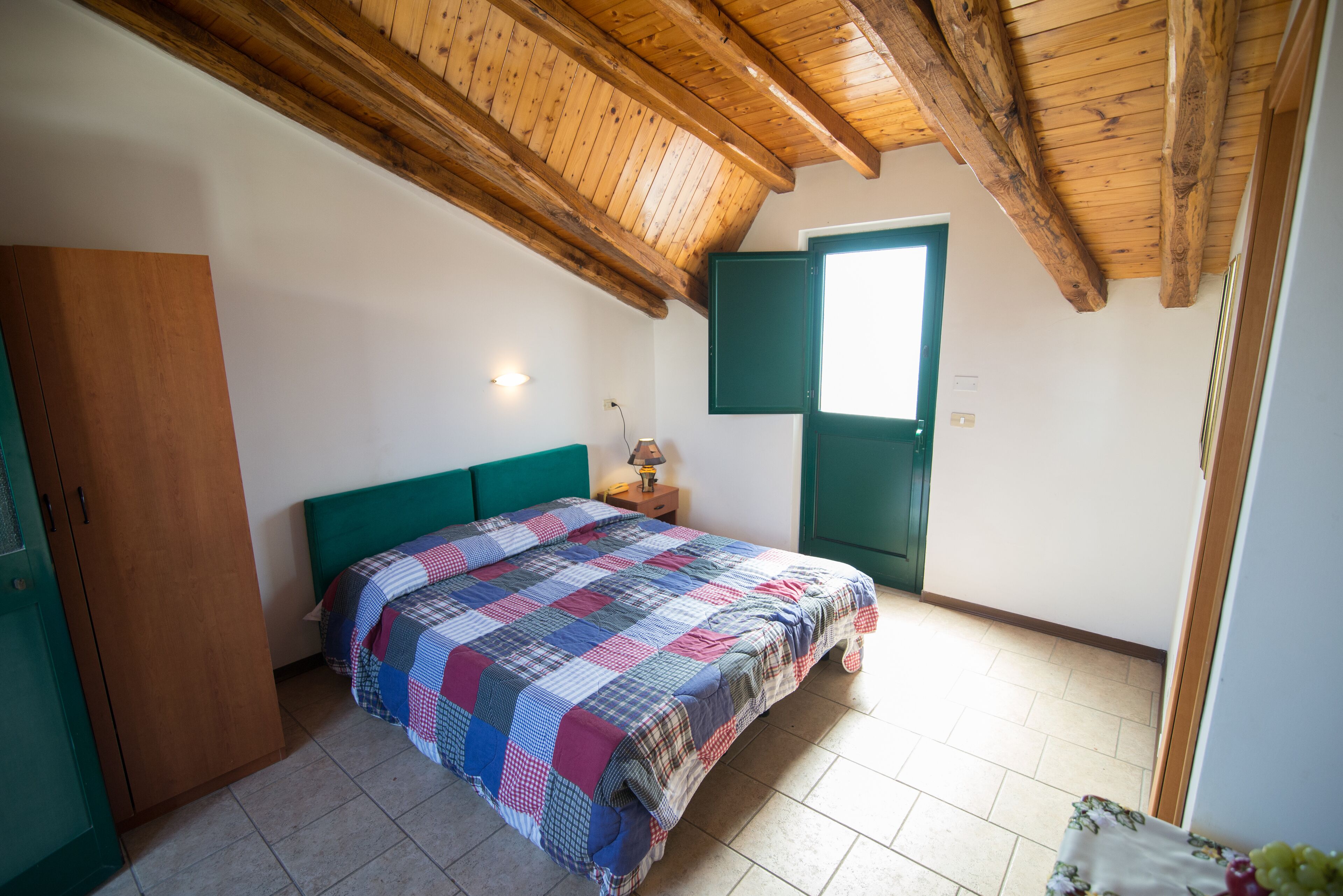 A bedroom with a wooden ceiling, a bed with a checkered bedspread, a green headboard, a nightstand with a lamp, and a door with a green frame.