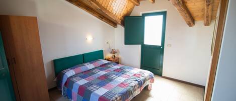 A bedroom with a wooden ceiling, a bed with a checkered bedspread, a green headboard, a nightstand with a lamp, and a door with a green frame.