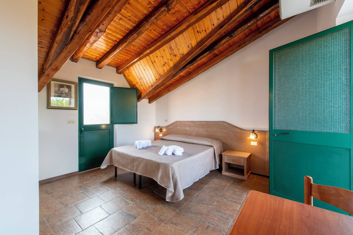 A bedroom with a wooden ceiling, a bed with a beige bedspread, a green door, and a small bedside table with a lamp.