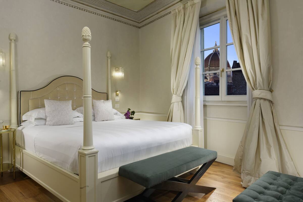 A neatly arranged bedroom with a four-poster bed, a bench, and a window with curtains.