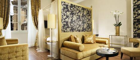 A luxurious bedroom with a four-poster bed, a sitting area with a sofa and armchairs, a round coffee table, and a large window with curtains.