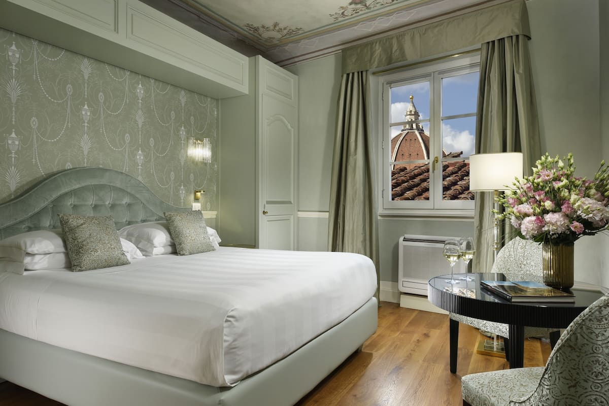 A neatly arranged bedroom with a large bed, a bedside table with a lamp and flowers, and a view of a building through a window.