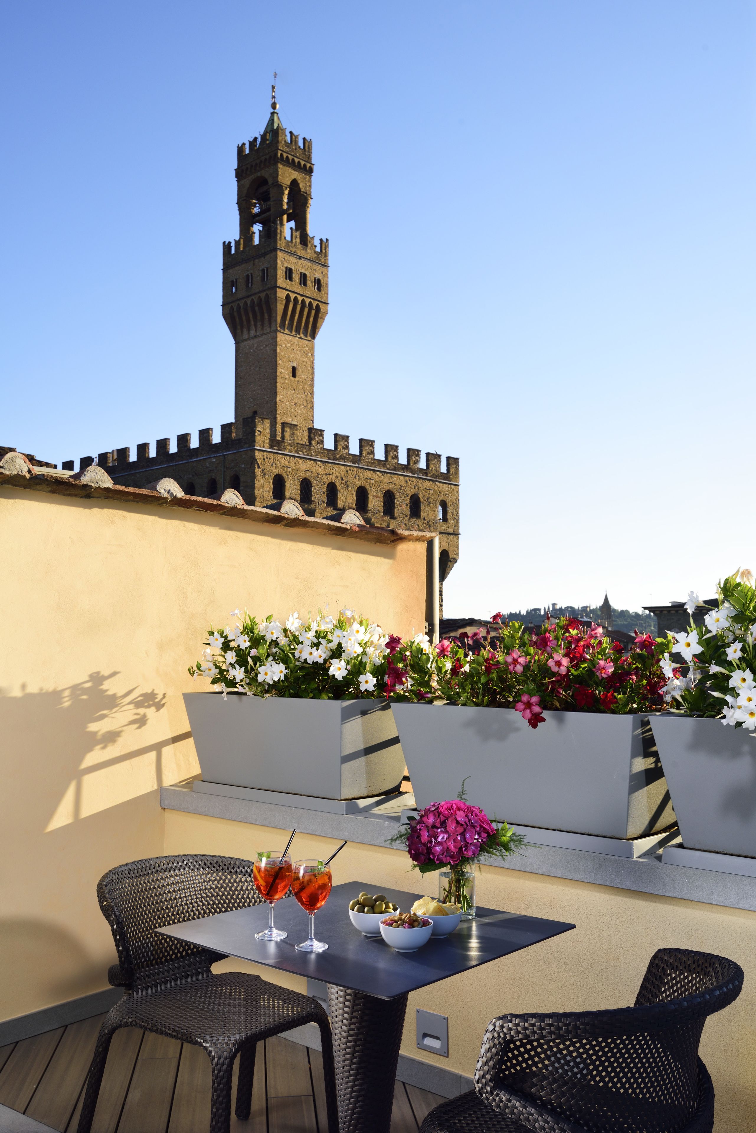 Eine Terrasse mit Tisch für zwei Personen, zwei Getränken, einer Oliven-Schale und einem Blumenstrauß. Im Vordergrund befindet sich ein großer Blumenkasten, im Hintergrund ist ein Burgturm sichtbar.