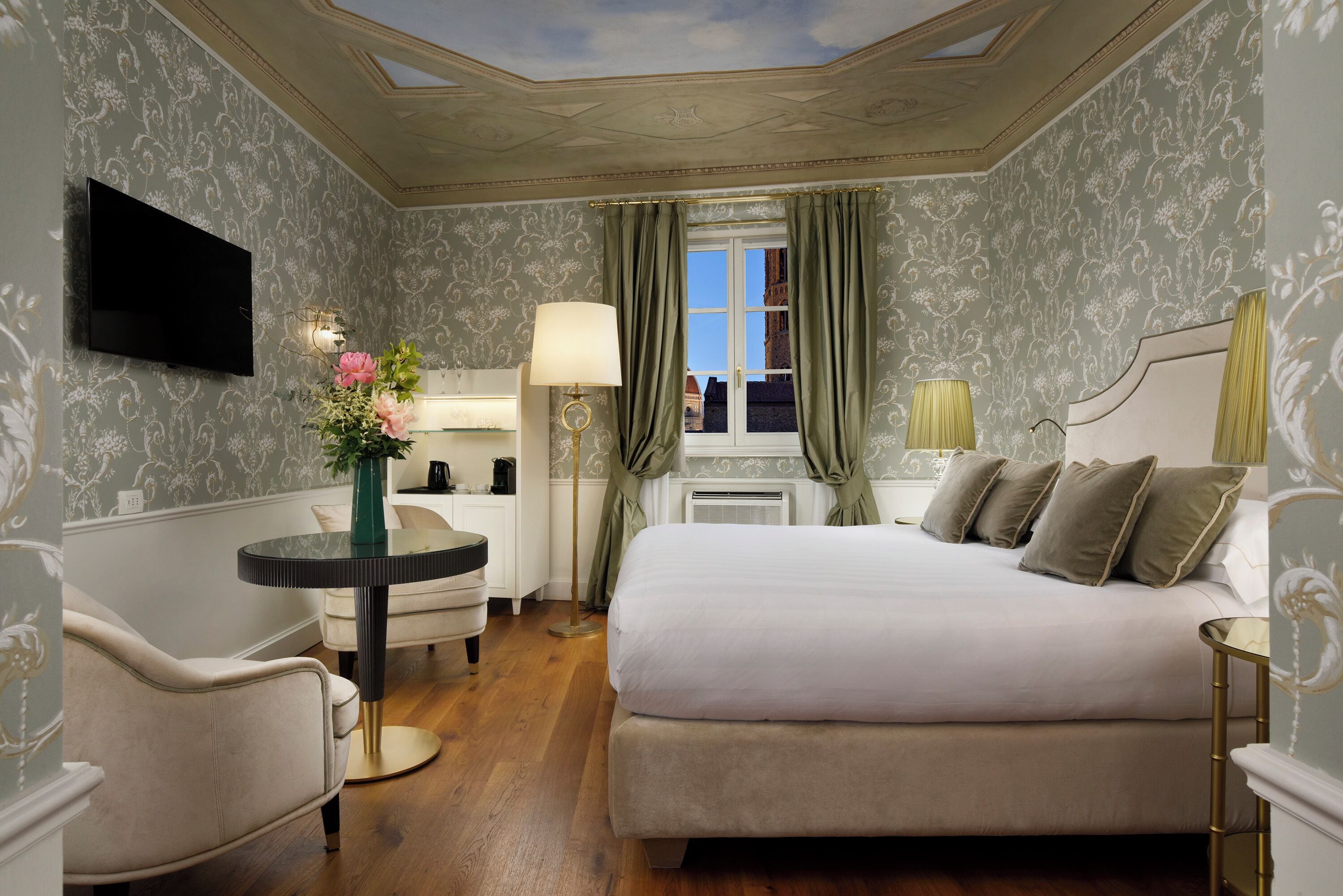 A hotel room with a large bed, a round table with a vase of flowers, a desk with a lamp, and a television mounted on the wall.