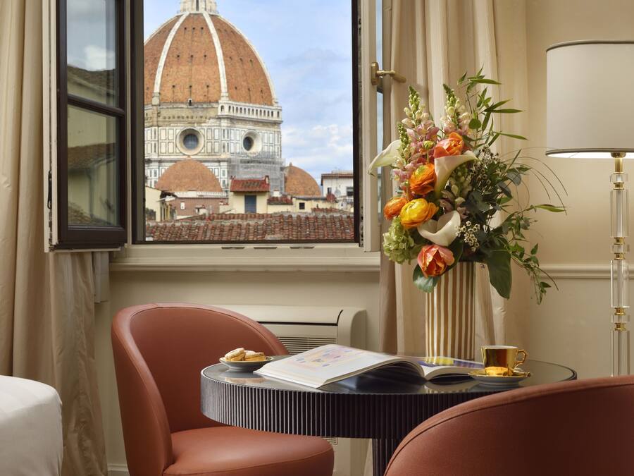 A hotel room with a view of a historic building, a vase of flowers, a table with a book and a cup, and a lamp.