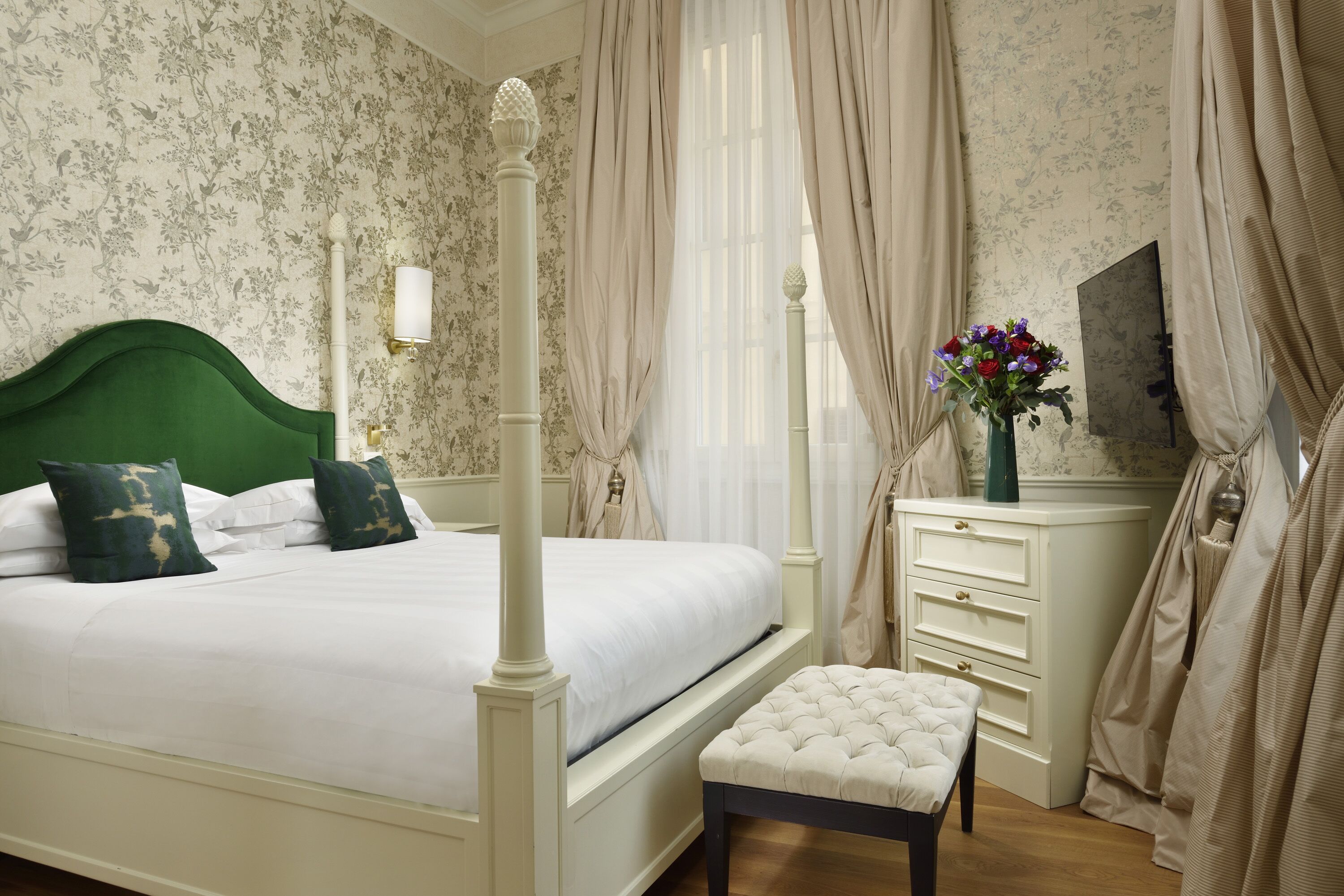 A four-poster bed with a green headboard, a bedside table with drawers, a mirror, and a vase with flowers.