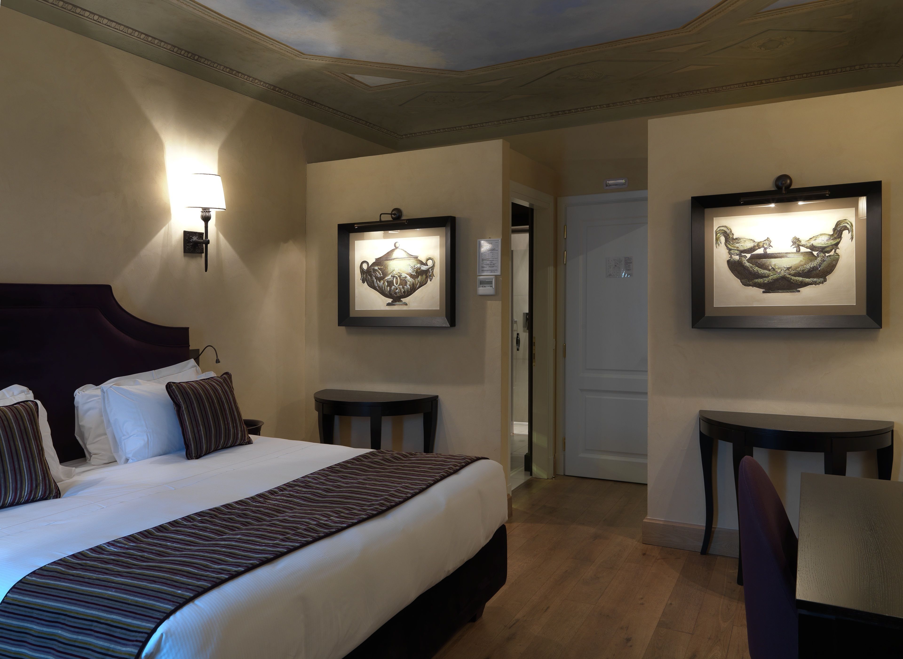 A hotel room with a large bed, bedside tables, a desk, and framed artwork on the walls.