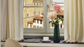 Ein Zimmer mit Fensterblick auf historische Gebäude, eine Vase mit Blumen, ein Sessel und ein kleiner Tisch mit einer Tasse.