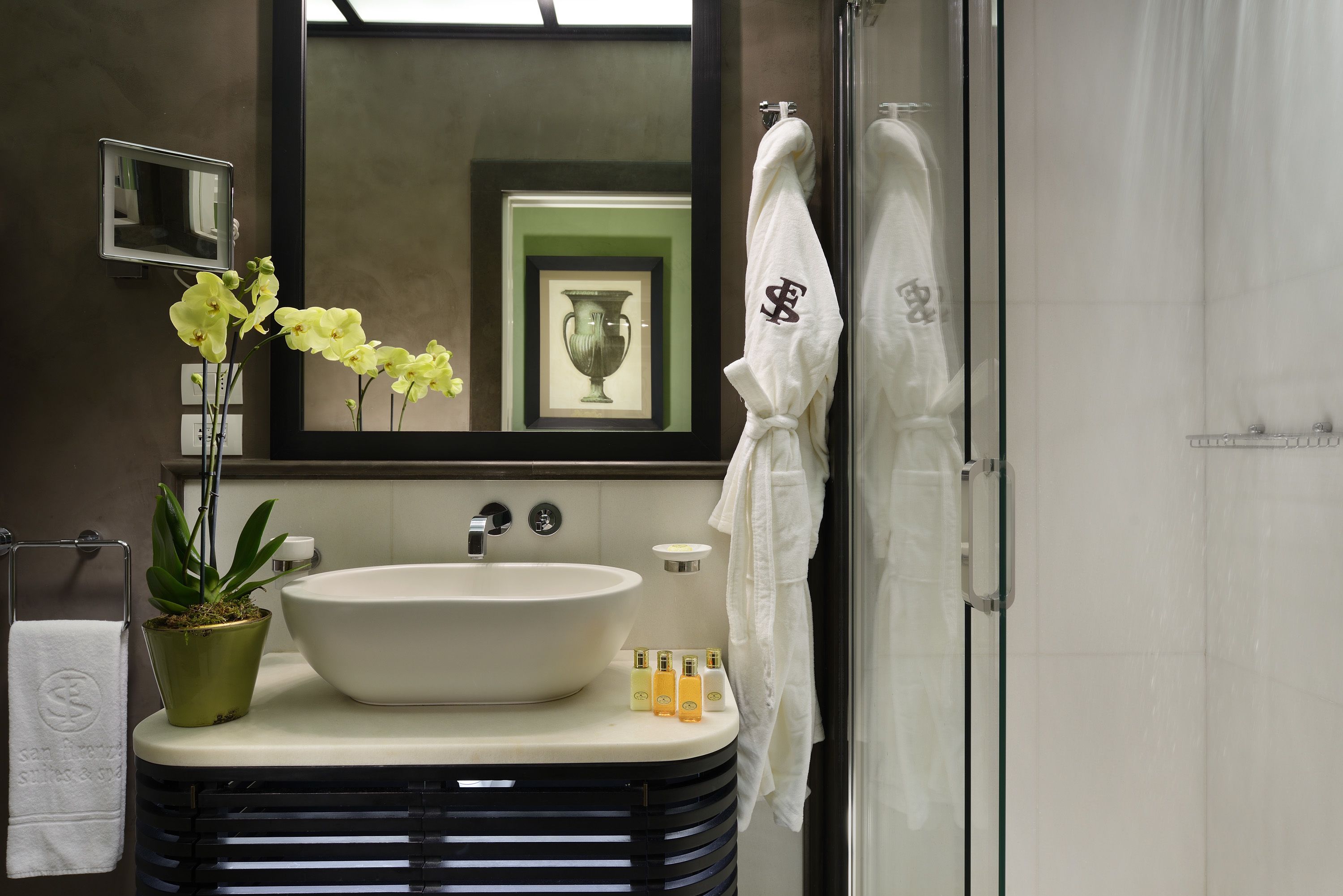 A bathroom with a large white sink, a potted orchid, a framed vase, and a towel rack with a white towel.