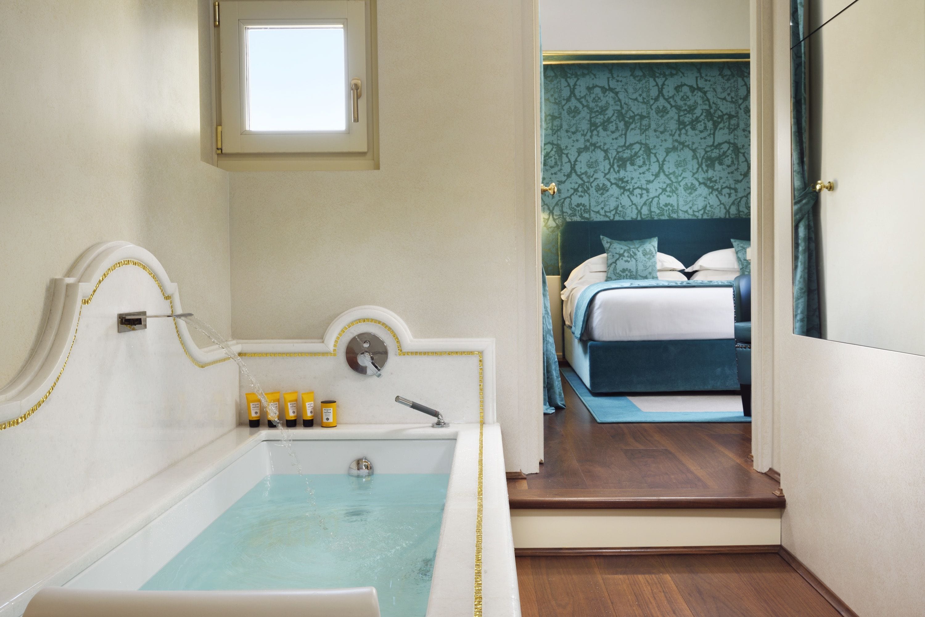 Un baño con bañera, una ventana y una puerta que da a un dormitorio con cama y mesita de noche.