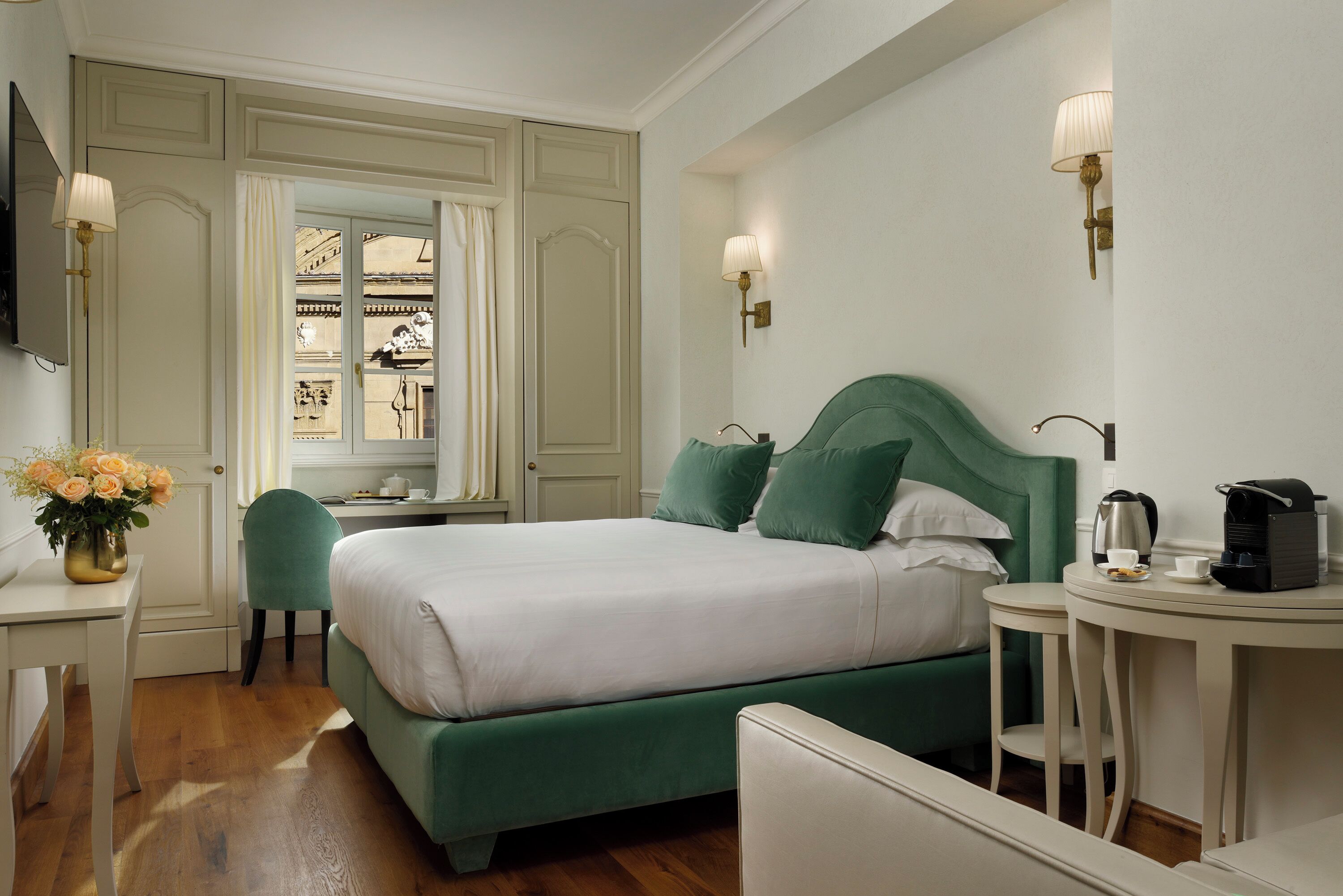 A neatly arranged hotel room with a bed, a desk, a chair, and a small table with a kettle and cup.