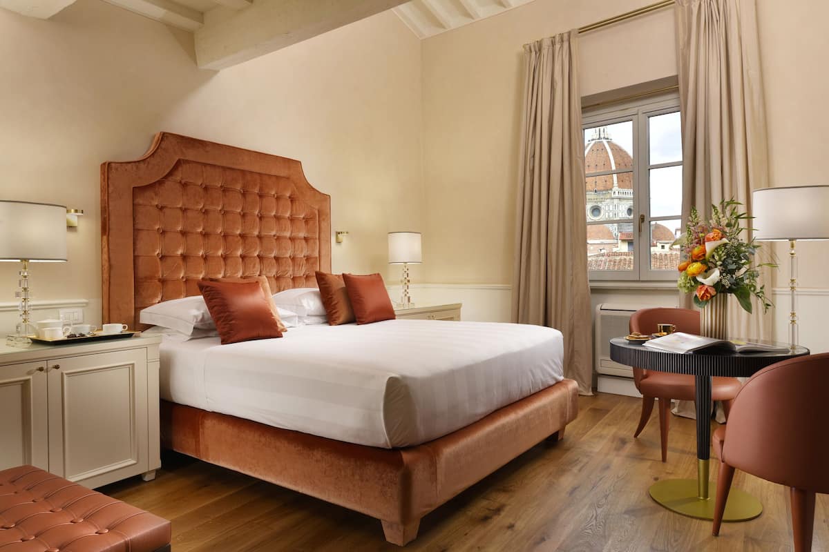 A hotel room with a large bed, a desk with a lamp, a chair, and a view of a building through a window.