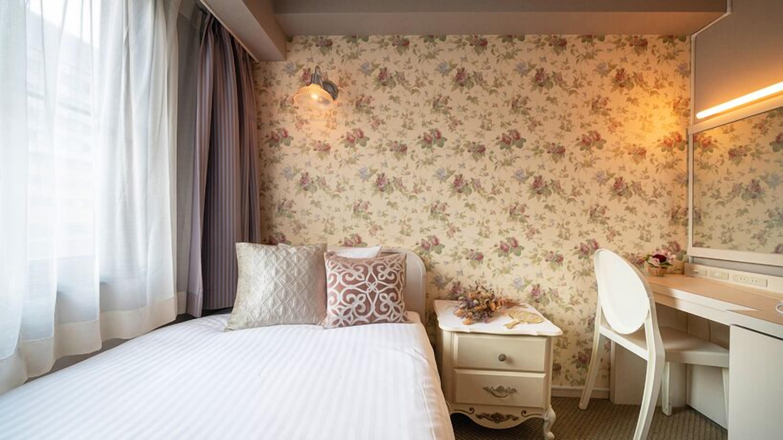 A hotel room with a bed, a desk, a chair, a nightstand, and a mirror.