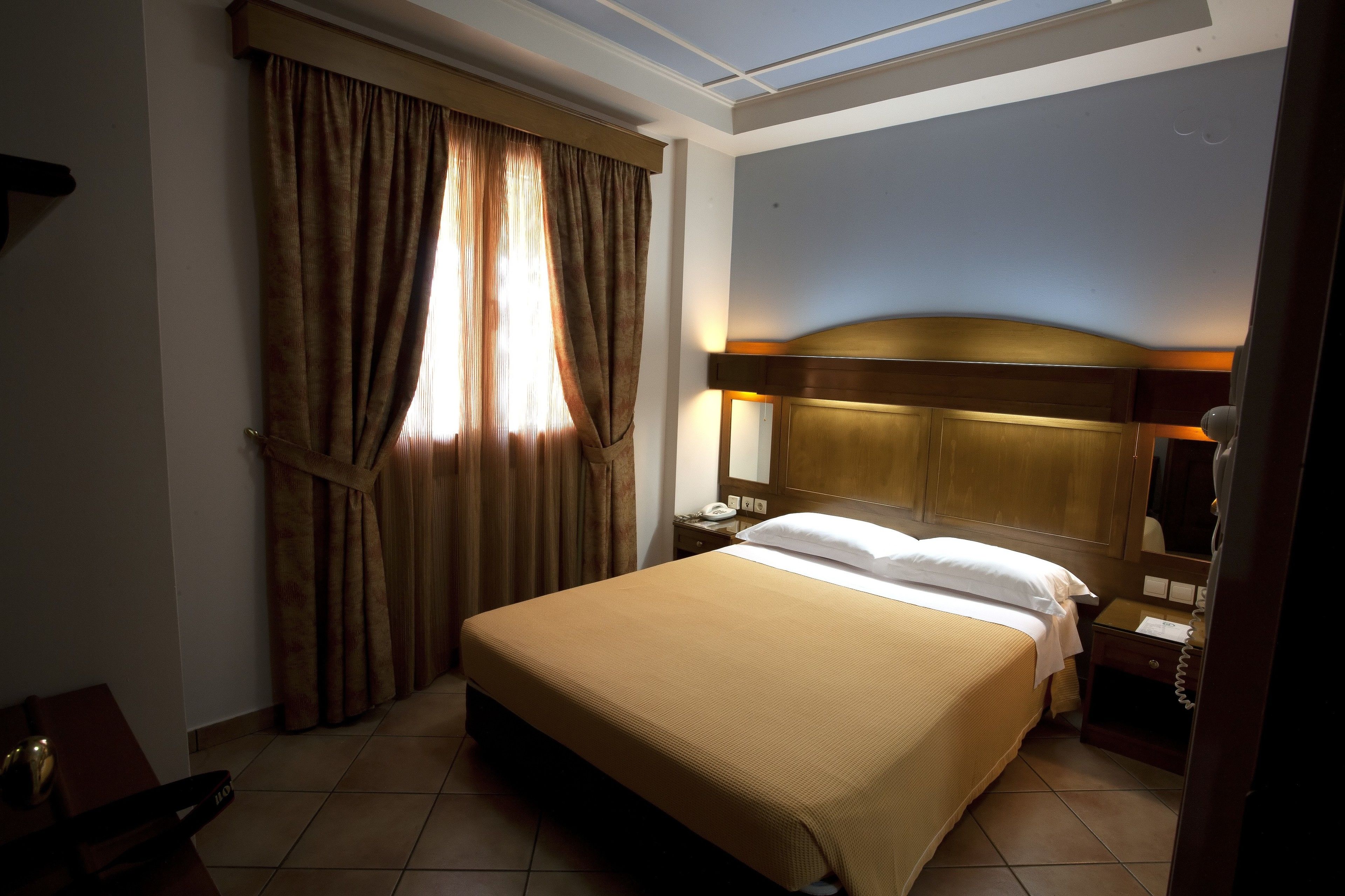 A hotel room with a large bed, a nightstand with a lamp, a wooden headboard, and a window with curtains.