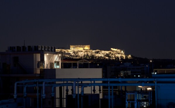 A nighttime view of a cityscape with a prominent ancient amphitheater illuminated on a hill, modern buildings in the foreground, and a body of water with a bridge. - Hotel Katerina (Athens)