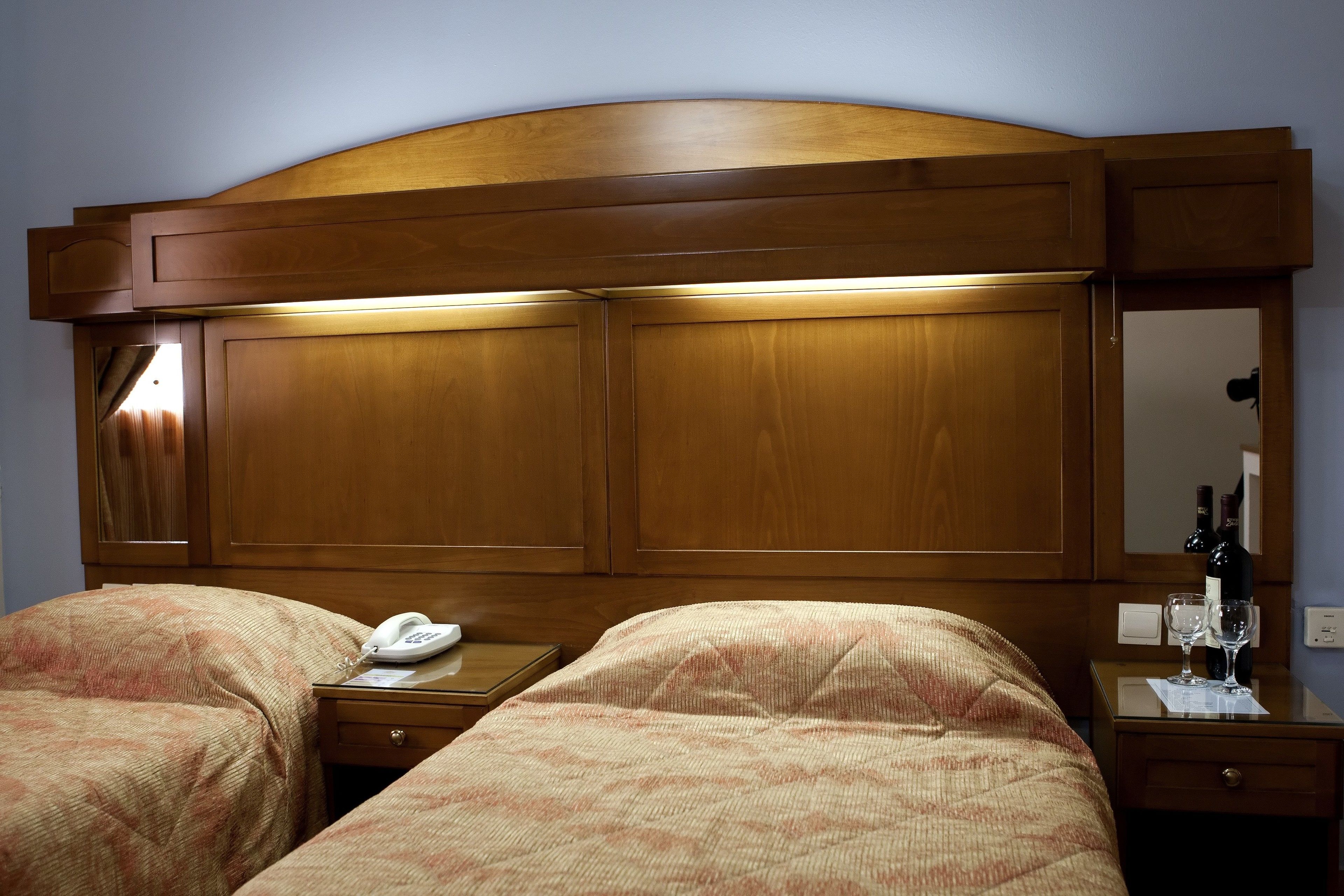 A hotel room with two beds, a wooden headboard, a bedside table with a phone, a mirror, and a small table with a bottle and glasses.
