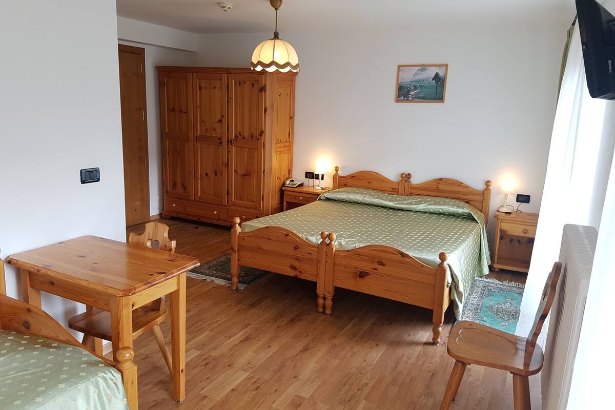 A wooden bed with a green bedspread, a wooden nightstand with a lamp, a wooden wardrobe, and a wooden table with a chair.
