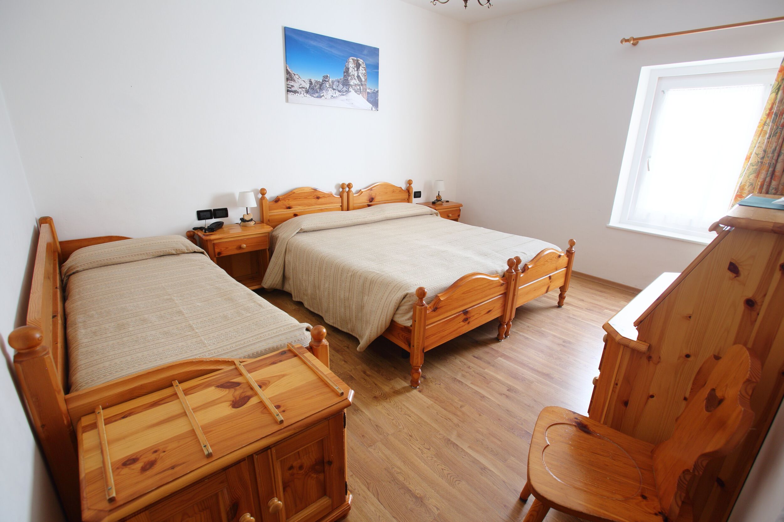 Une pièce avec deux lits en bois, une chaise en bois, une table de chevet avec une lampe et une fenêtre avec des rideaux.