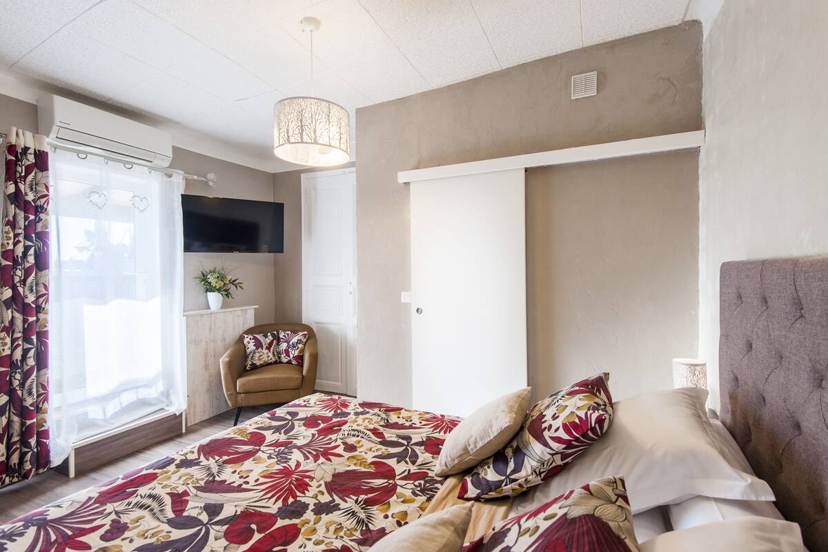 Quarto com cama, uma cadeira, televisão e janela com cortinas.