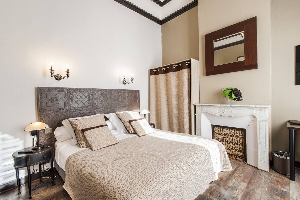 Um quarto bem arrumado com uma cama grande, uma mesa de cabeceira com abajur, uma lareira e um espelho na parede.
