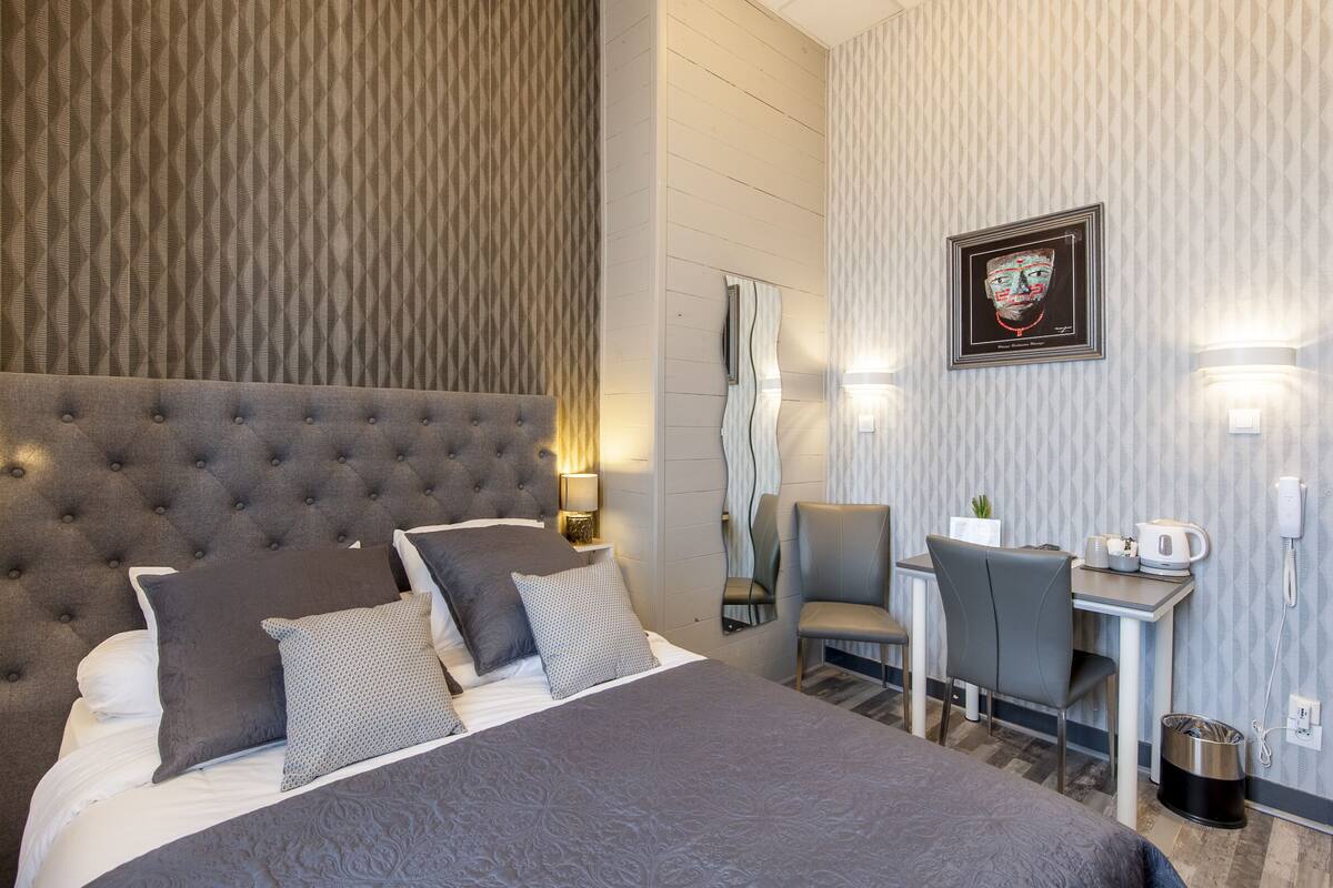 Quarto de hotel com cabeceira estofada, cama com roupa de cama cinzenta, escrivaninha com cadeira, uma obra de arte emoldurada na parede e uma pequena planta em vaso.