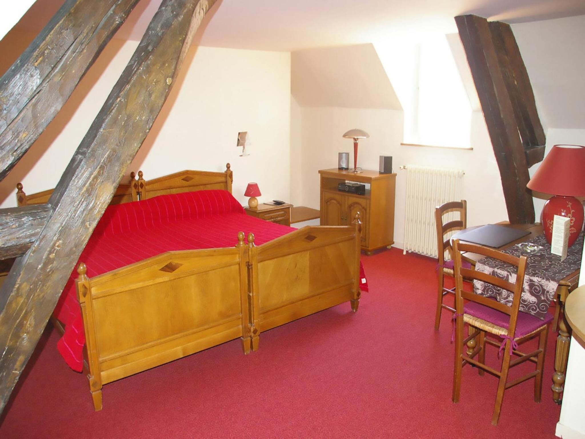 A bedroom with a wooden bed, a red carpet, a desk with a chair, a radiator, and a window with a view of the sky.