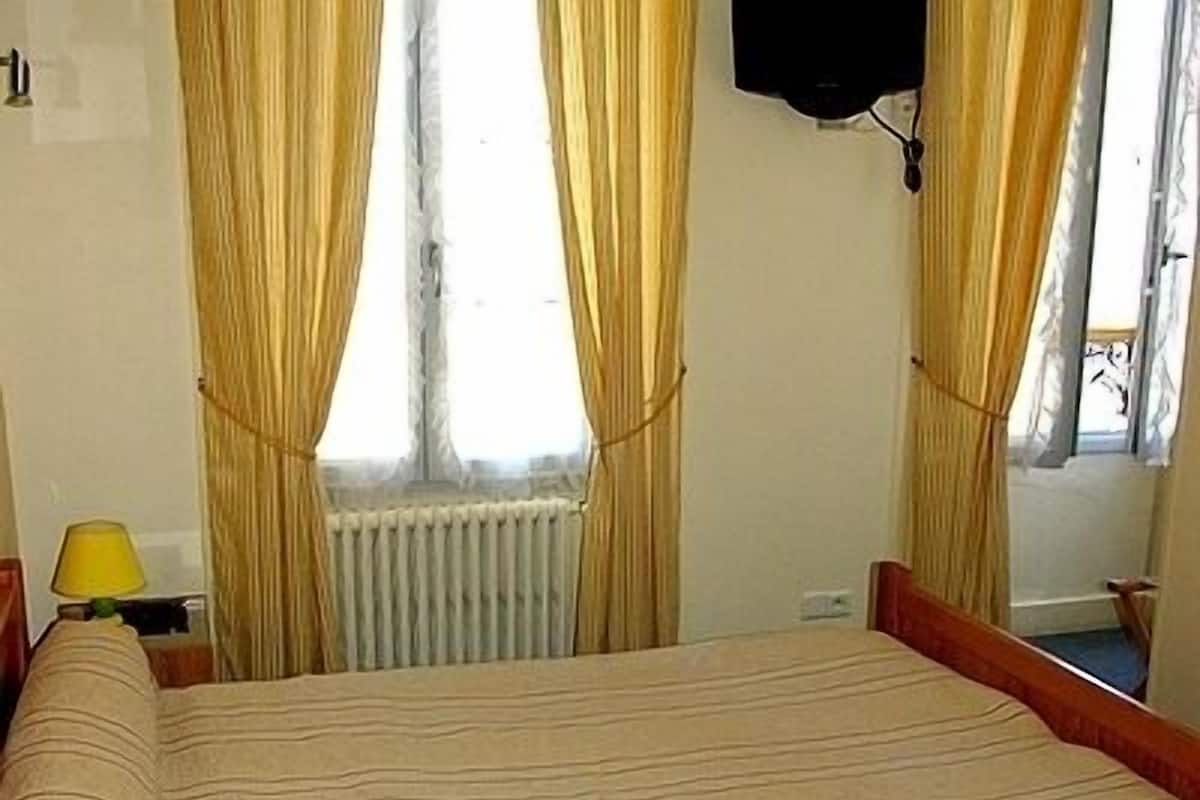 Een hotelkamer met een bed, een raam met gordijnen, een radiator, een lamp en een spiegel.