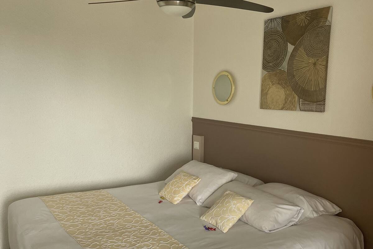 Uma cama bem arrumada com colcha estampada e travesseiros, um ventilador de teto e um espelho fixado na parede.