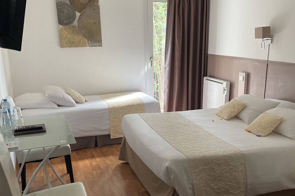 Quarto de hotel com duas camas de solteiro, uma mesa com cadeira, televisão presa na parede e janela com cortinas.