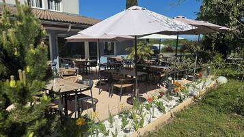 Un espace extérieur aménagé en terrasse, avec des tables et des chaises, entouré d’un jardin où poussent différentes plantes et fleurs.