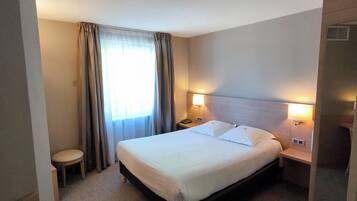 Hotellihuone, jossa on sänky, yöpöydällä olevat lamput, yöpöytä, tuoli ja ikkuna, jossa on verhot.