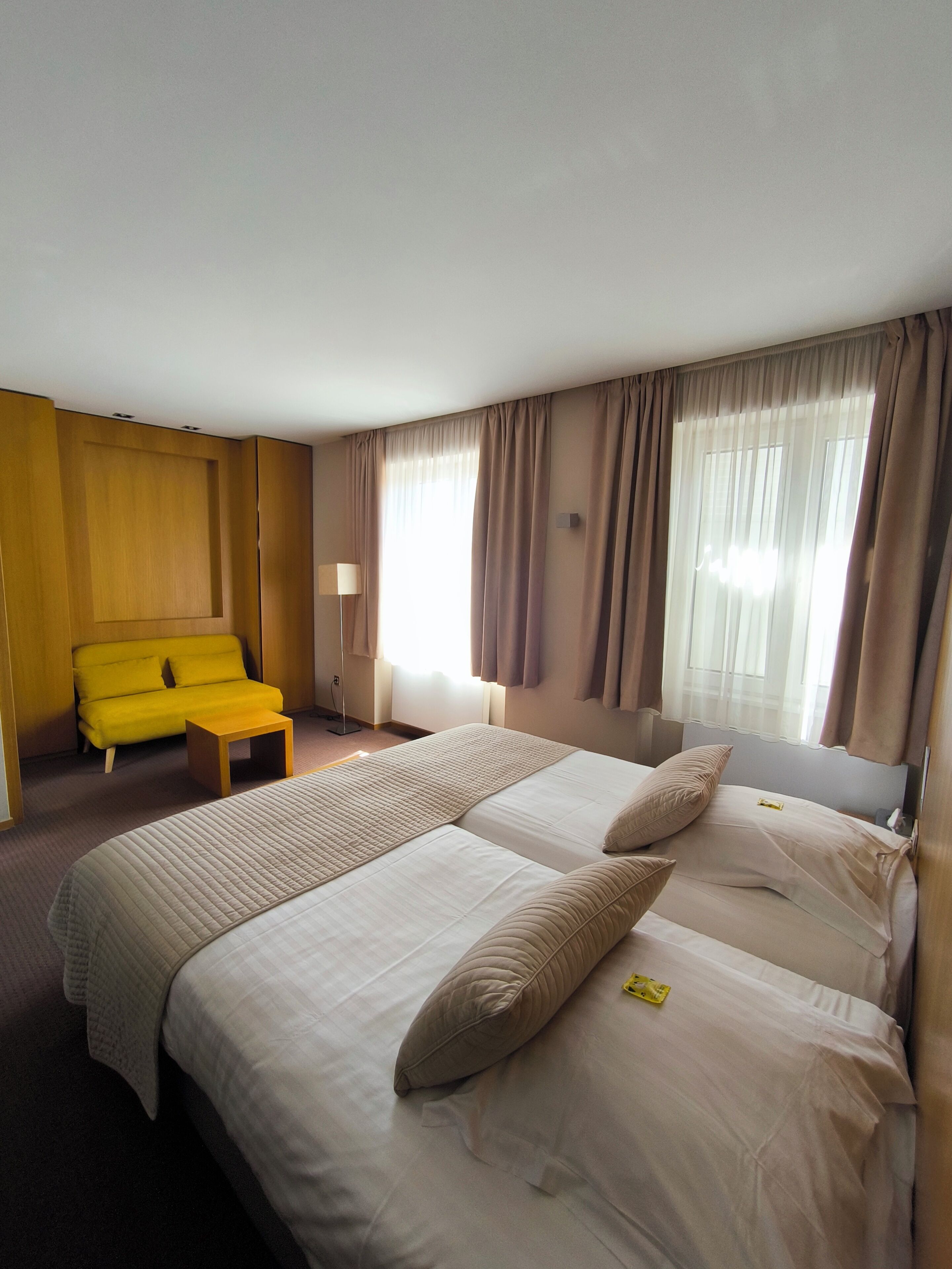 Una habitación de hotel con una cama, un sillón amarillo, una mesita y una ventana con cortinas.