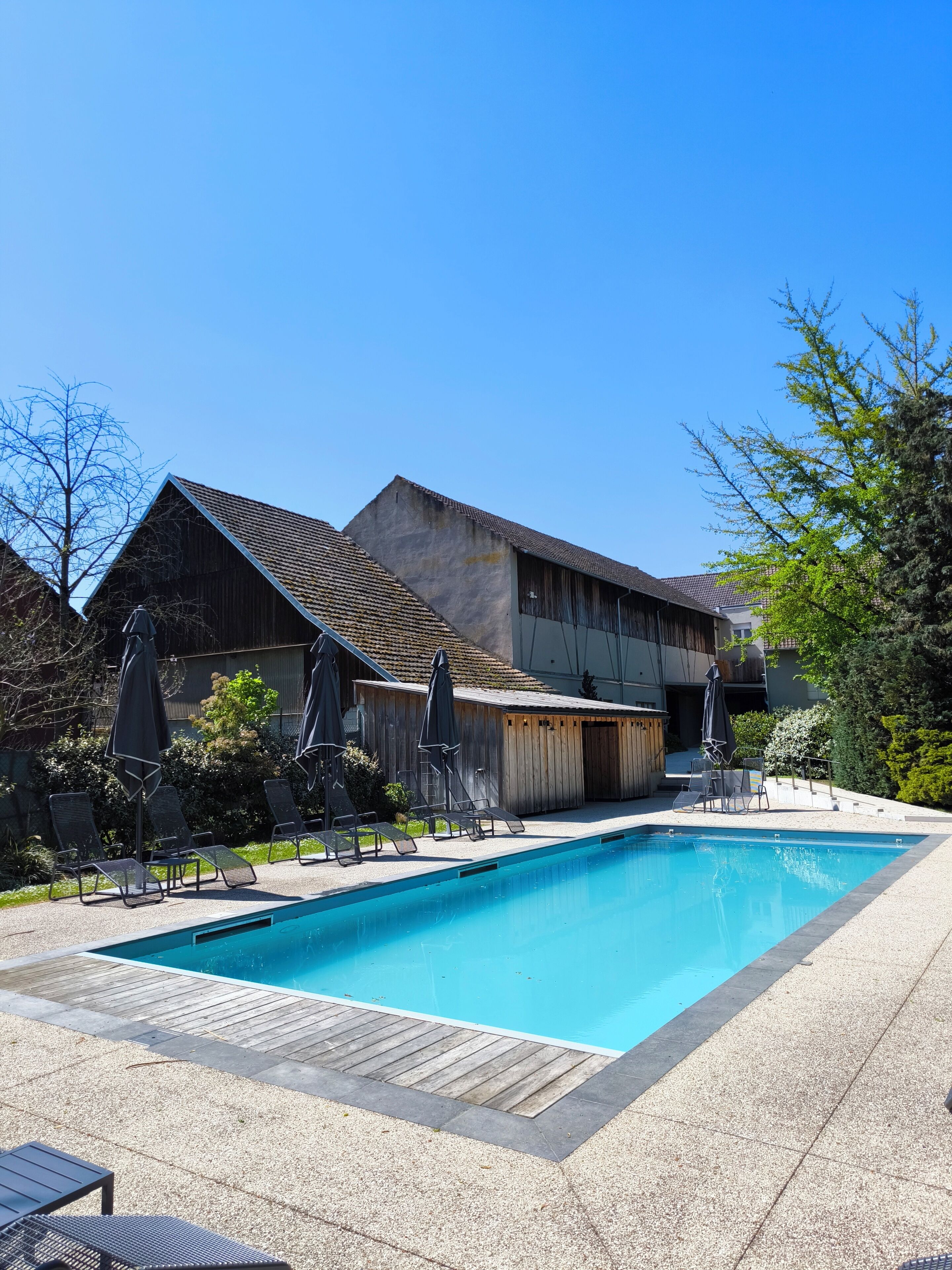Una piscina con lettini e ombrelloni, un edificio in legno e un cielo azzurro limpido.