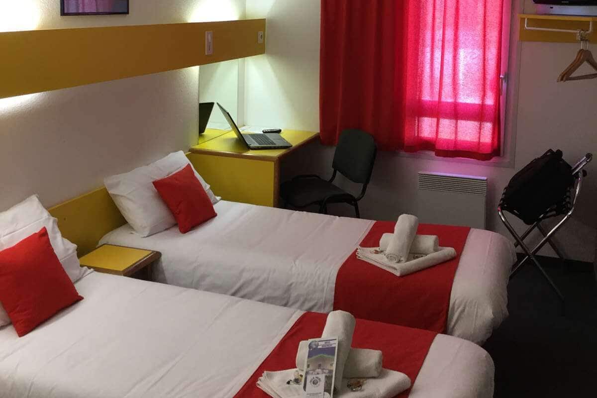 A hotel room with two beds, a desk with a laptop, a TV, and a red curtain.