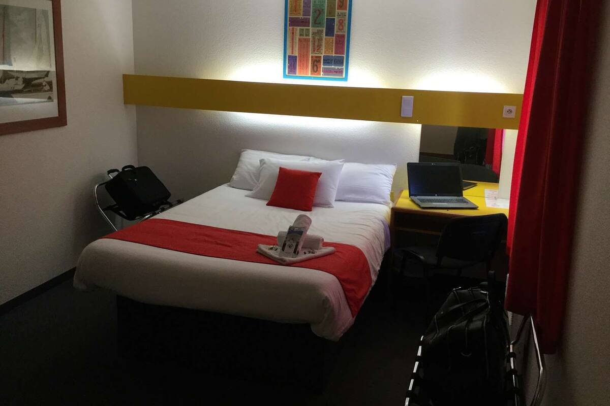 A hotel room with a bed, a desk with a laptop, a chair, a suitcase, and a wall-mounted shelf with a colorful picture.