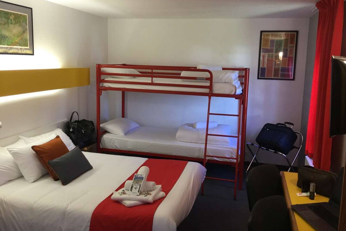 A hotel room with a bunk bed, a single bed, a desk with a chair, a laptop, and a small table with a bottle and a tissue.