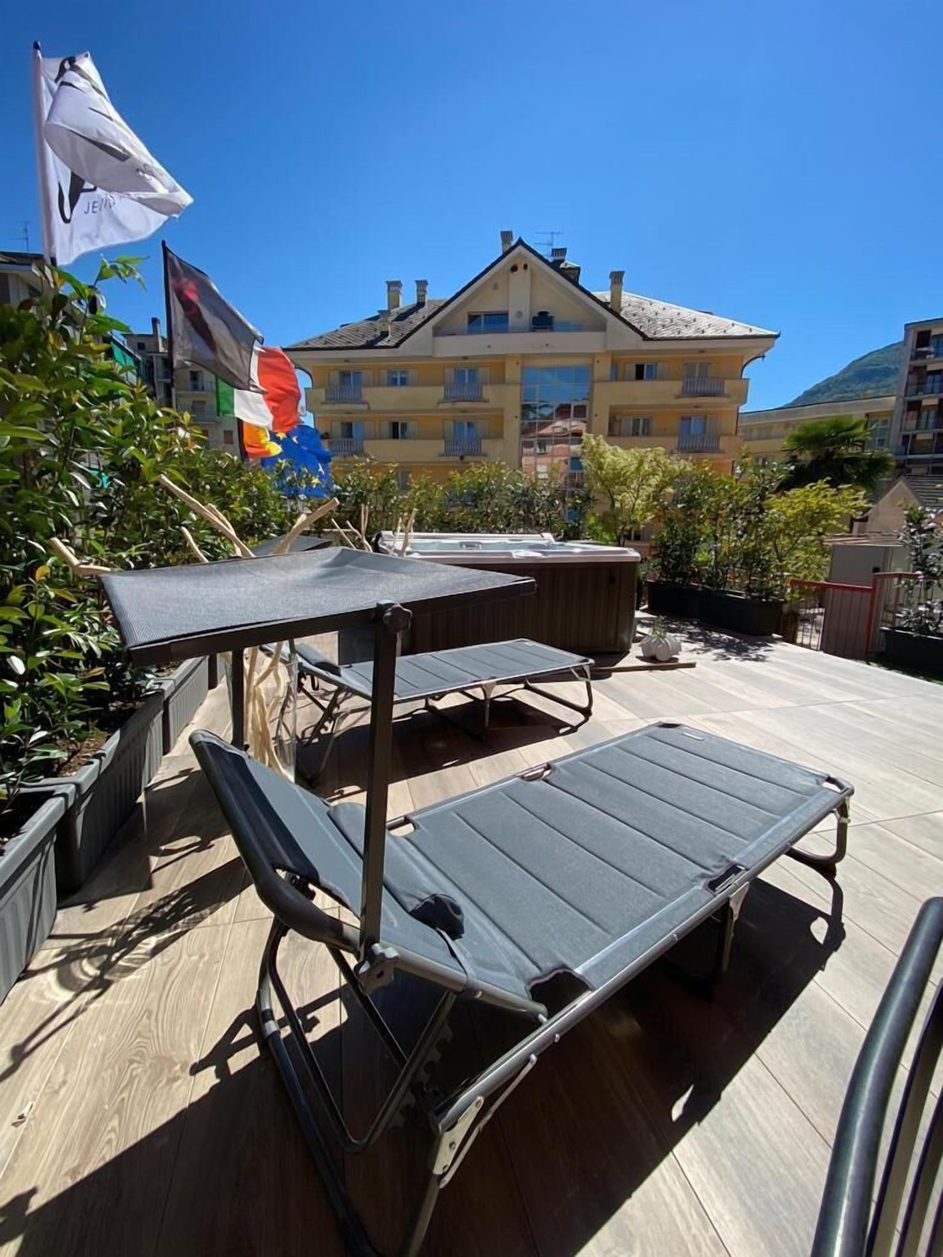 Terraza en la azotea con una mesa y sillones de descanso, rodeada de plantas en macetas y banderas.