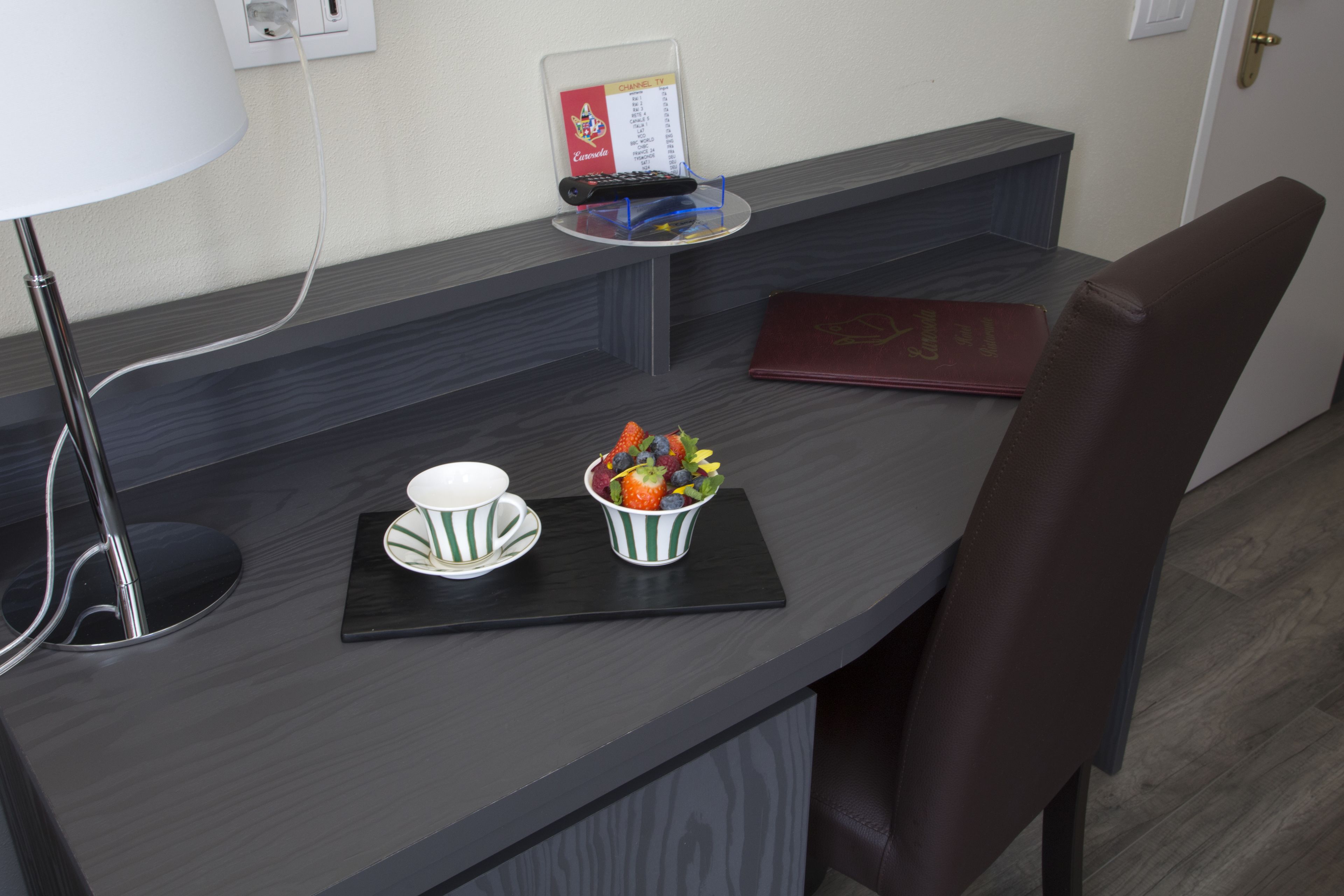 A modern hotel room with a dark wood desk, a remote control, a menu card, a bowl of fruit, and a red book.